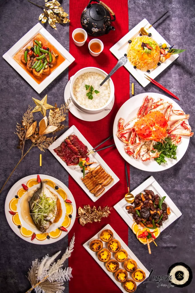 A table set with a variety of dishes including stir-fried vegetables, white rice in a bowl, crab, fried rice, dim sum, roast duck, braised meat, fish, and egg tarts. There's a black teapot with two cups, and decorative golden elements on the table.