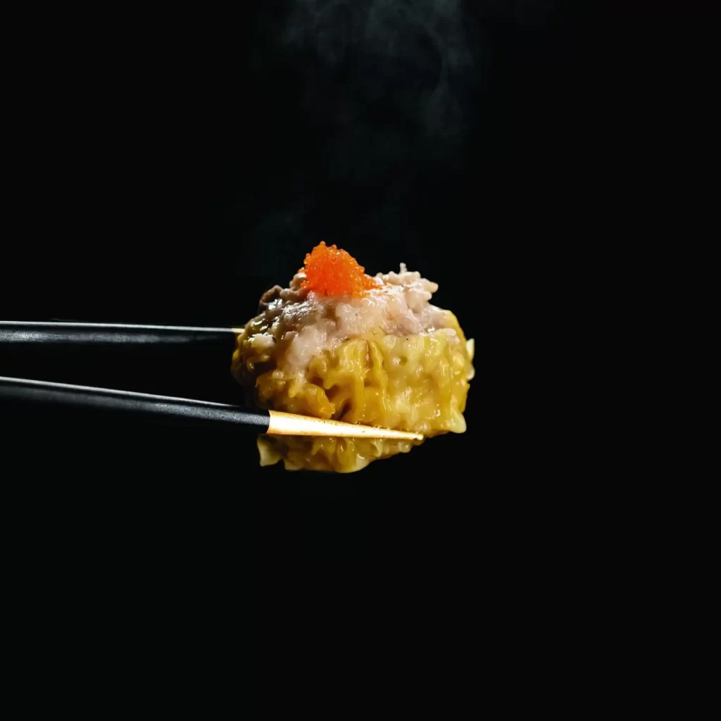 A pair of chopsticks is holding a piece of food against a black background. The food appears to be a dumpling topped with a small amount of orange roe and a portion of minced meat. Steam is rising from the dumpling, indicating that it is hot.