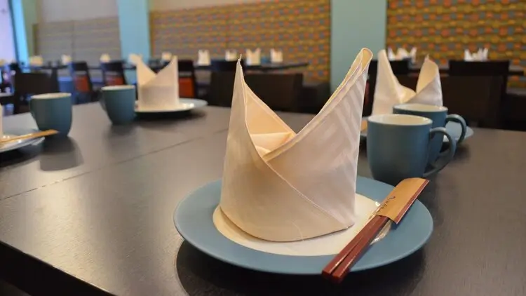 A close-up of a dining table set with blue plates, white folded napkins resembling crowns, teal mugs, and silverware placed on the plates. The background shows more similarly set tables with a modern decor featuring patterned walls and seating.